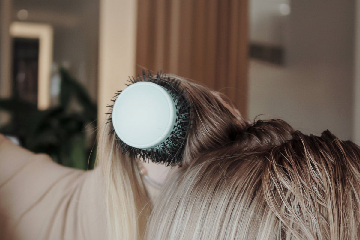 A close-up lifestyle shot of a person using a round hairbrush to detangle or style their light brown and blonde hair.